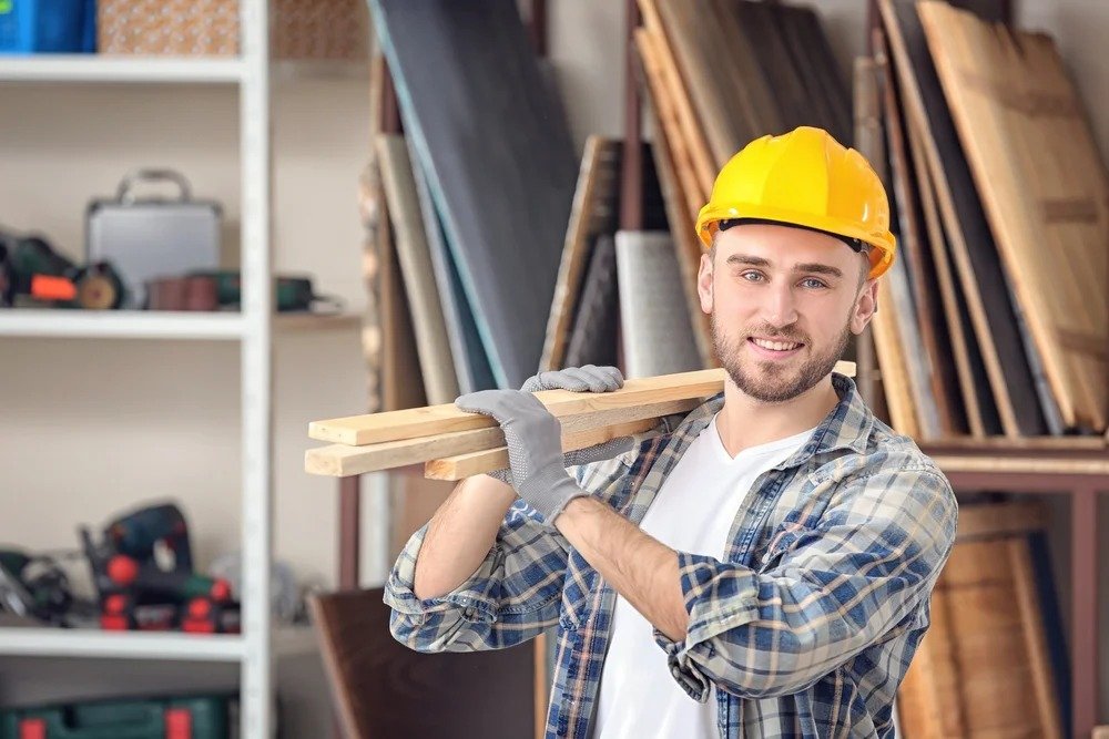 A man wearing a hard hat holds a wooden plank, standing in a construction environment.
